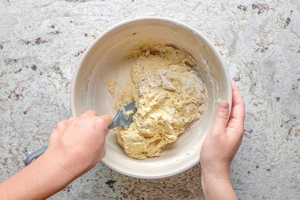 overhead shot of A person mixing thick dough in a white bowl with a spatula on a light, textured countertop