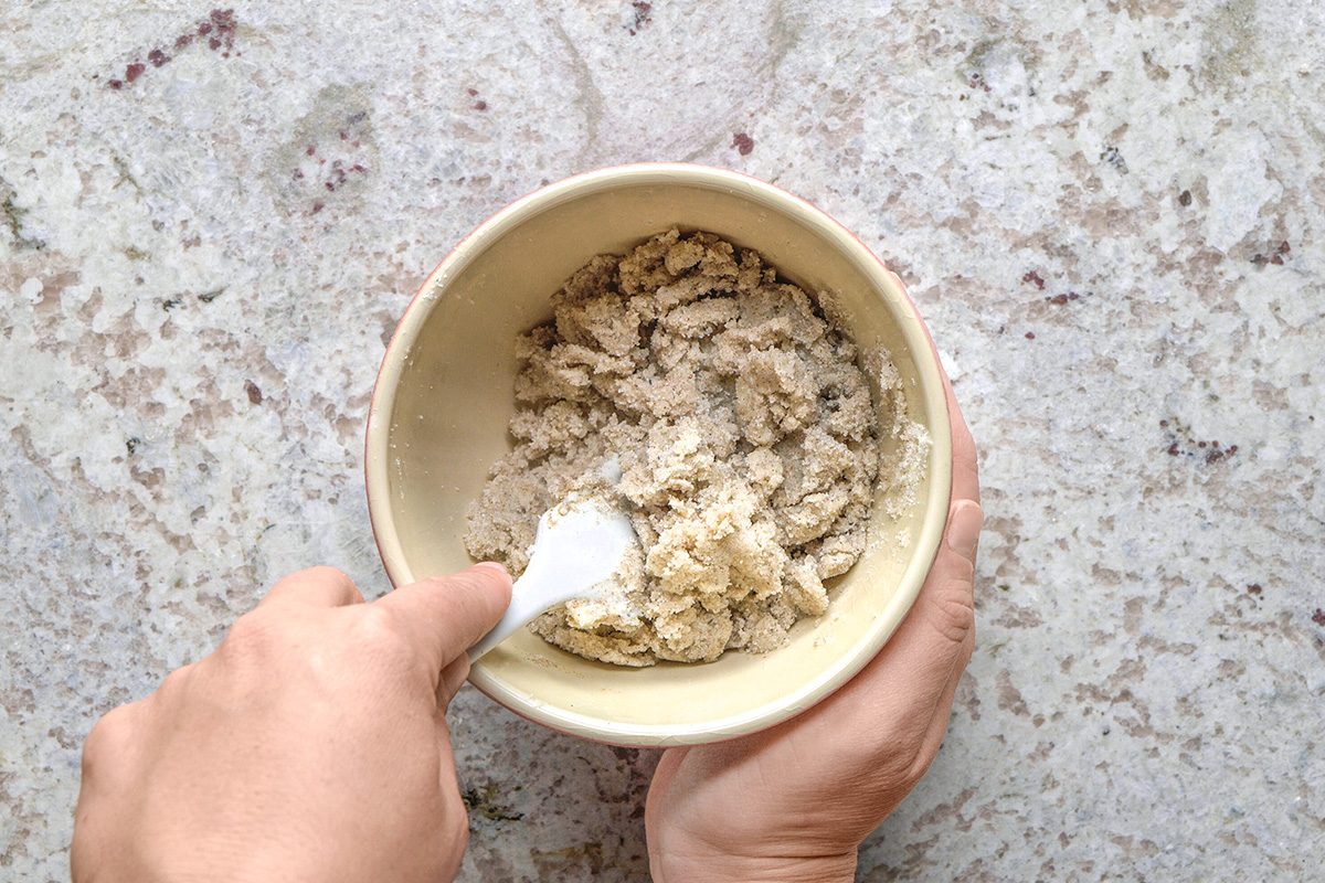overheda shot of A person holds a bowl and mixes a crumbly, light brown dough or mixture with a white spatula on a light speckled countertop