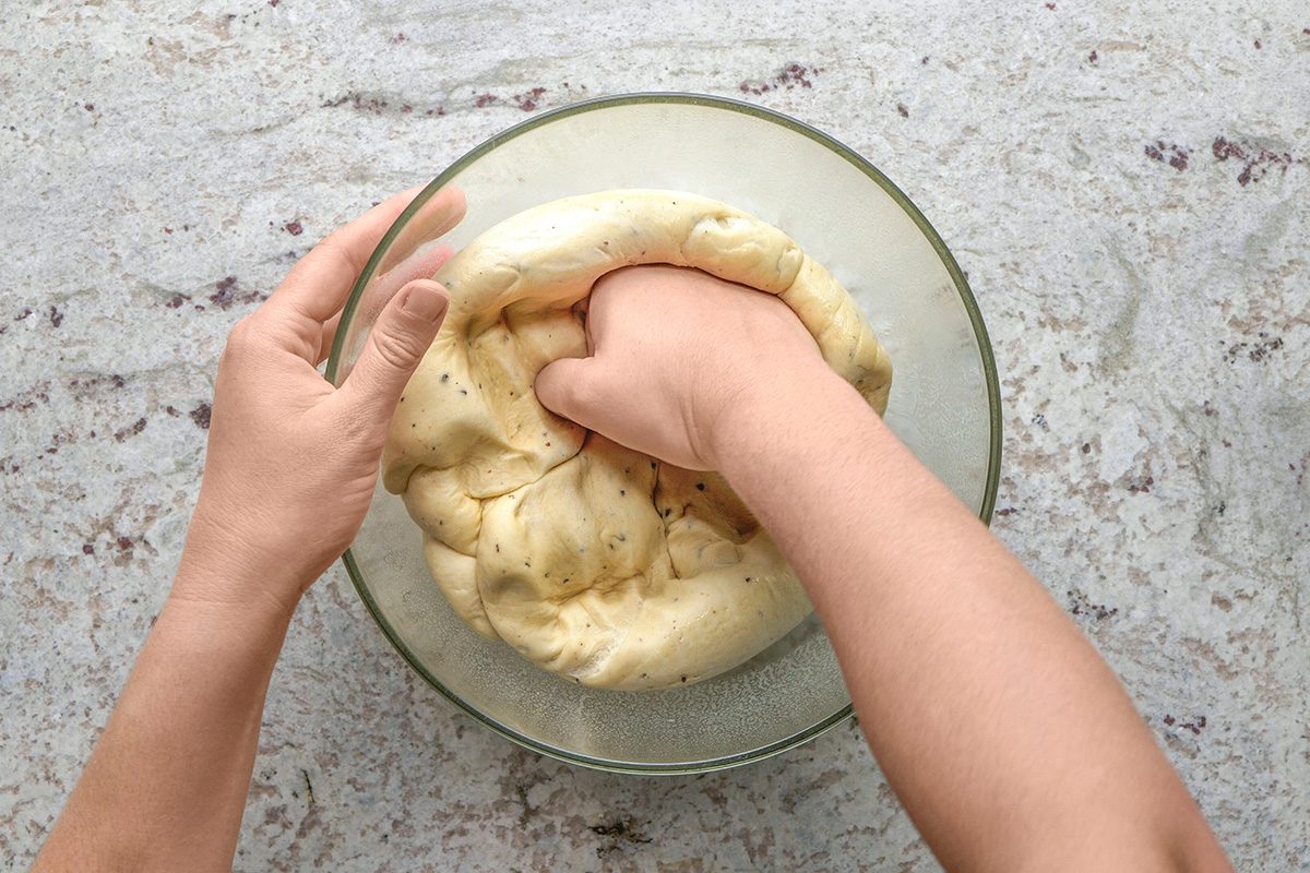 overhead shot of A person’s hands knead dough in a clear glass bowl on a light colored countertop; The dough appears to be soft and elastic, and one hand is pressing into it while the other steadies the bowl