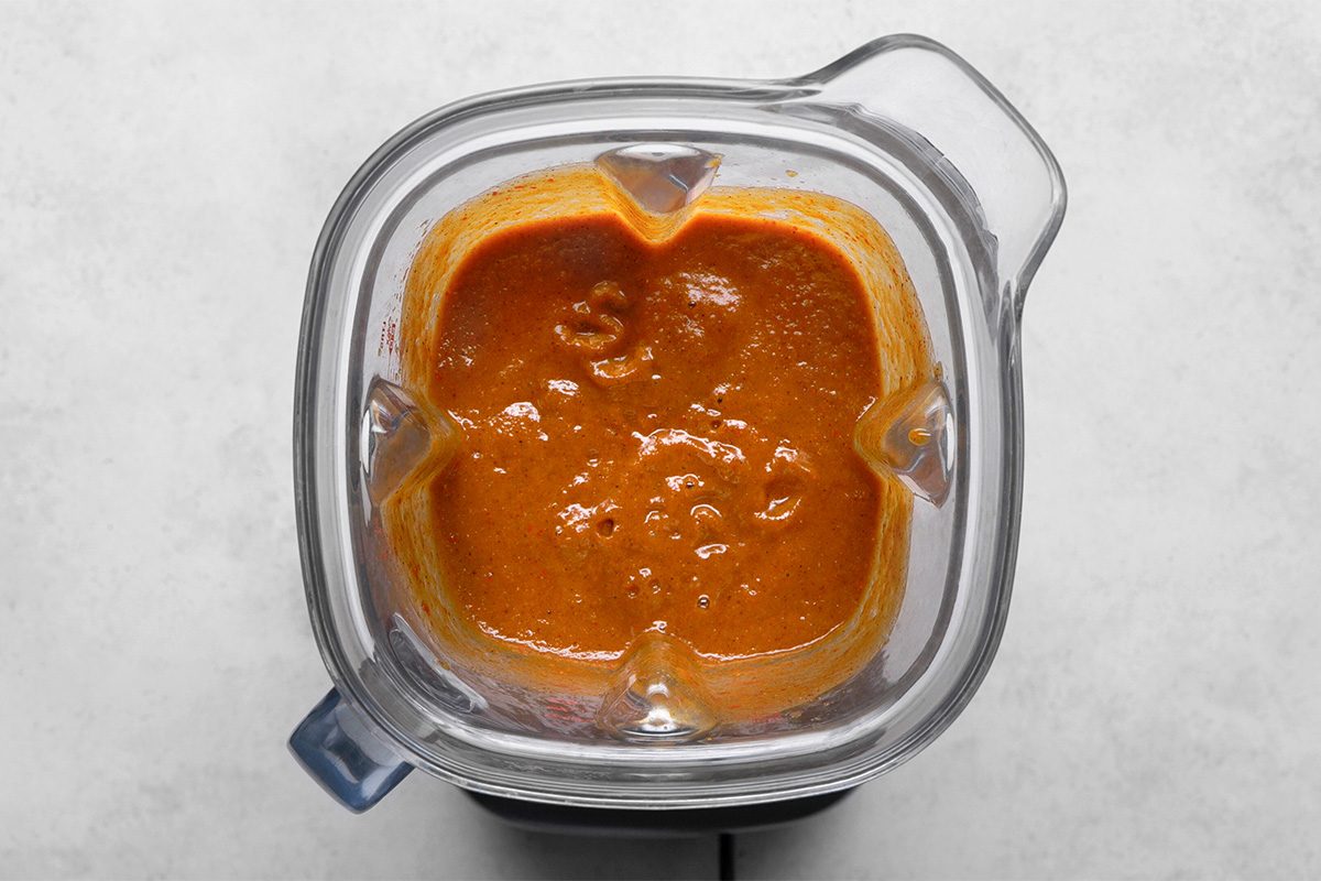 Overhead view of a blender filled with a smooth, orange-red sauce against a light-colored background.