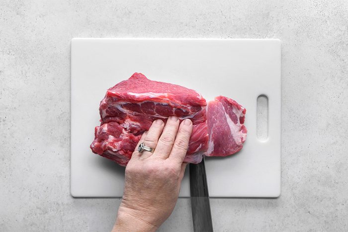 A hand holds a raw pork roast on a white cutting board, while a knife is used to cut through the meat.