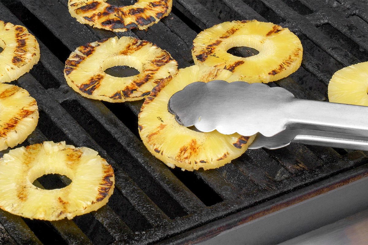 Metal tongs hold a ring of pineapple on a grill with several other pineapple rings, showing grill marks on the fruit.