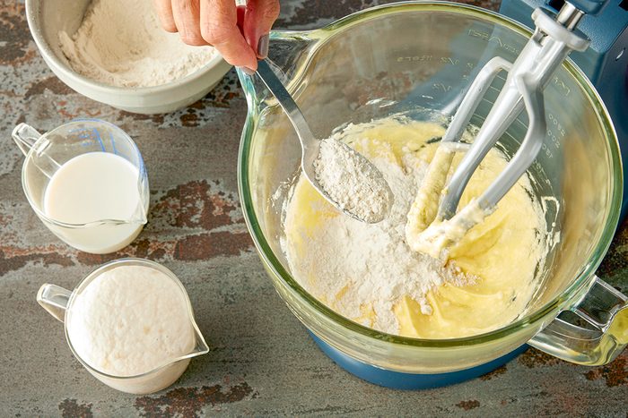 Overhead shot of a hand adding flour with a spoon into a mixing bowl of batter beside a stand mixer; nearby sit a bowl of flour; a measuring cup of milk; and a cup with a foamy white mixture on a brown countertop