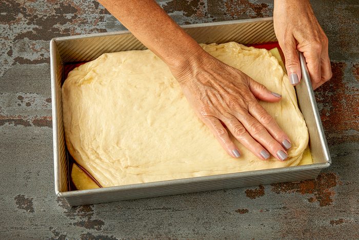 Overhead shot of a person pressing a sheet of dough evenly into a rectangular metal baking pan on a dark textured surface