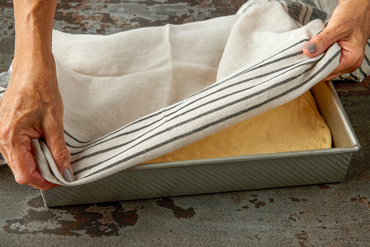 3/4 shot of hands lifting a striped kitchen towel to reveal bread dough rising in a rectangular metal baking pan on a dark countertop