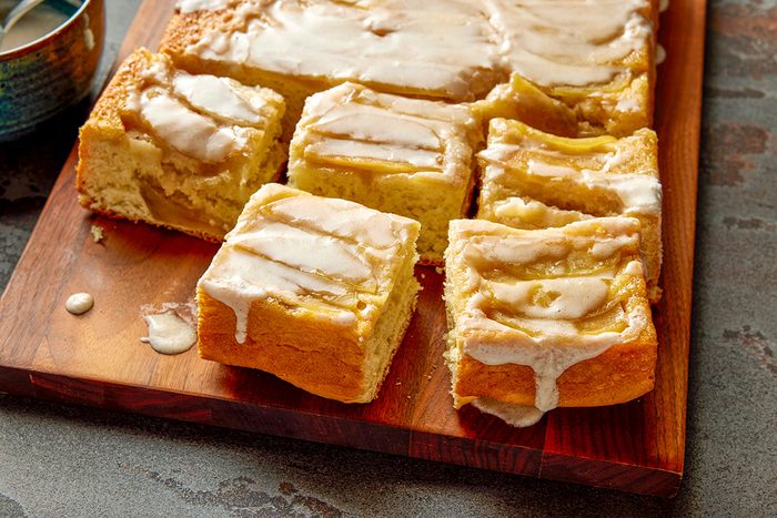3/4 shot of sliced apple bread topped with white glaze on a wooden cutting board; showing a golden crust and visible fruit pieces inside each moist square slice