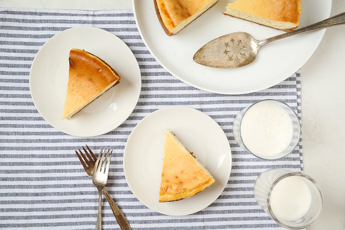 Three slices of cheesecake on white plates, a serving spatula on a platter, two glasses of milk, and two forks, all placed on a gray and white striped cloth.