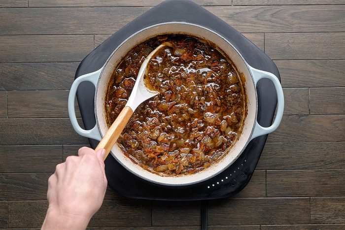 A hand stirs caramelized onions in a white pot on a black electric cooktop, set on a dark wooden surface.