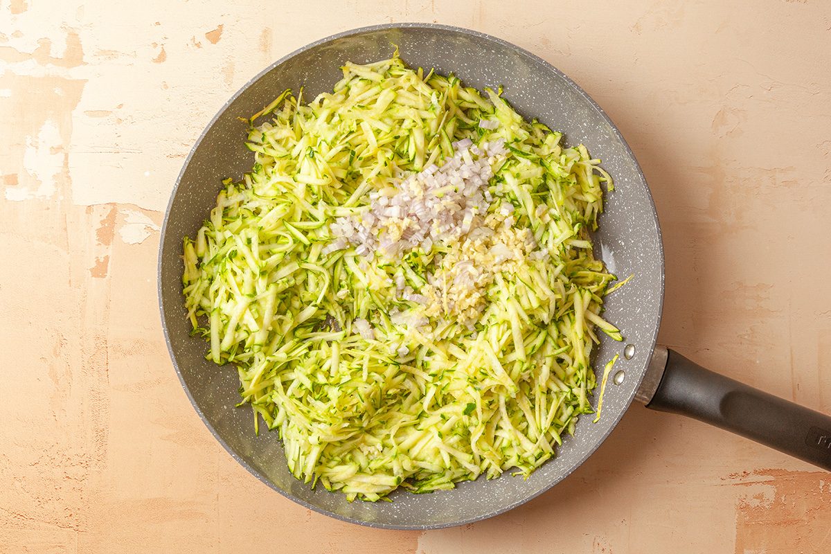 This is a close-up top-down shot; a frying pan filled with shredded zucchini; finely chopped onions; and minced garlic on a light background; ingredients appear fresh and uncooked