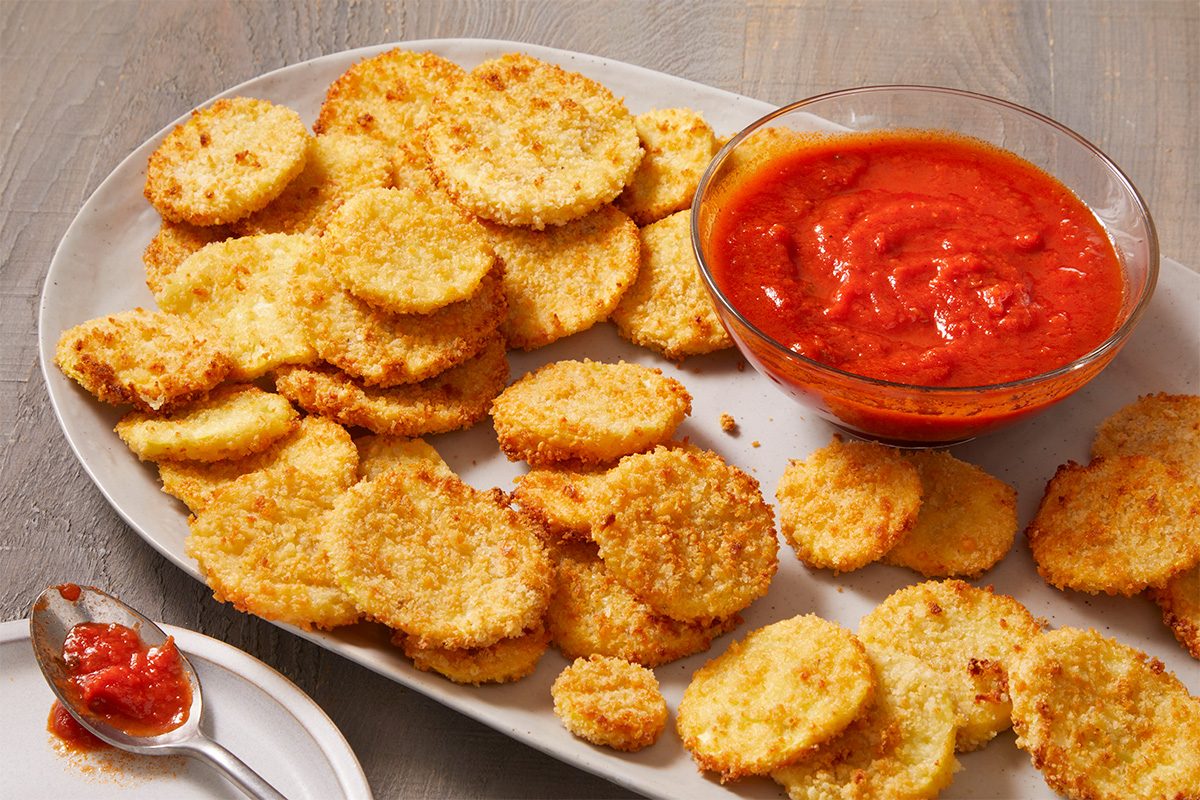 A platter of crispy breaded zucchini rounds surrounds a bowl of marinara sauce for dipping, with a spoonful of sauce resting nearby on the table.