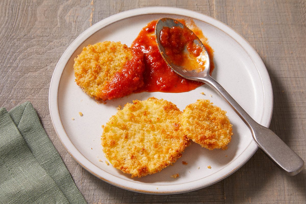 A white plate with three breaded, fried eggplant slices and a spoon resting in a pool of red marinara sauce, on a wooden table with a folded green napkin beside the plate.