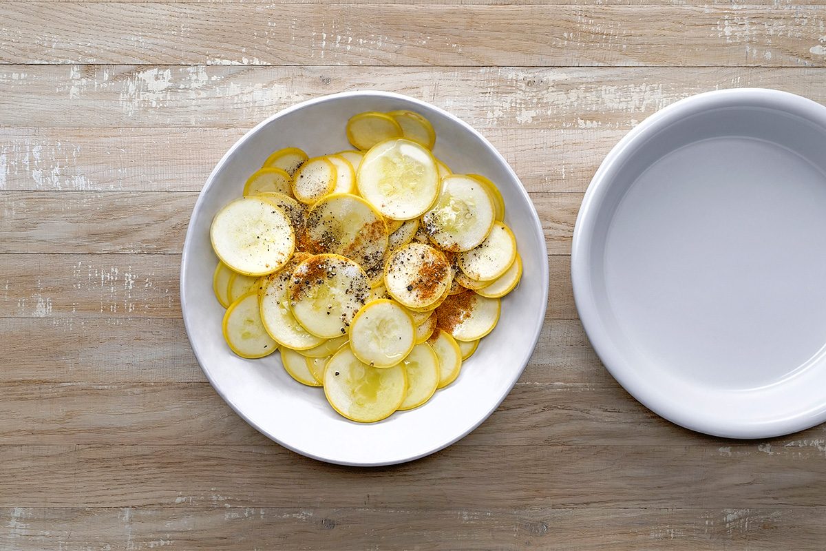 A white plate filled with thinly sliced yellow squash, seasoned with pepper and spices, sits on a wooden table next to an empty white bowl.