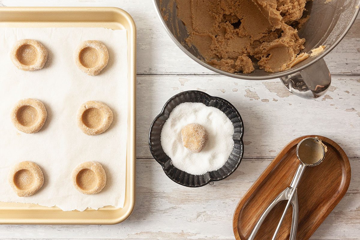 A baking tray with six unbaked cookies with thumbprints sits on parchment paper. Nearby are a bowl of cookie dough, a bowl of sugar with a dough ball, and a cookie scoop on a wooden tray.