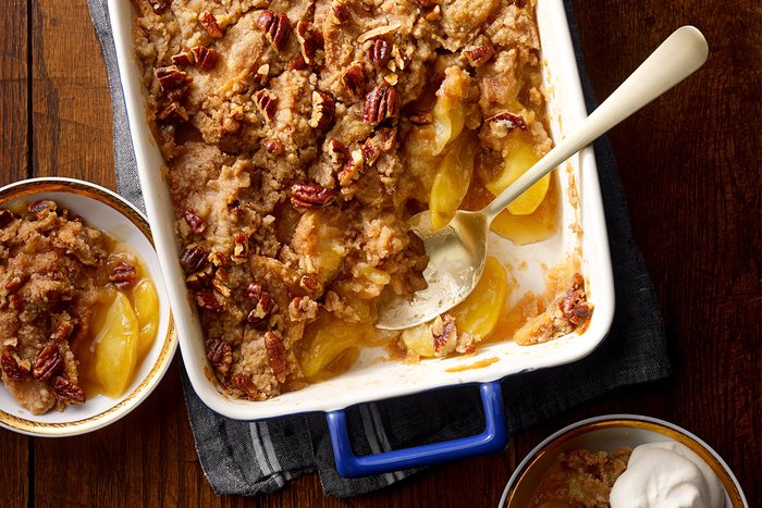 A baked apple crisp dessert in a white rectangular dish, topped with pecans and a crumbly streusel. A spoon rests inside, with servings in small bowls nearby on a wooden table.