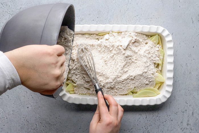 A person pours flour mixture from a gray bowl over sliced apples in a white baking dish while holding a whisk. The dish is on a light gray countertop.