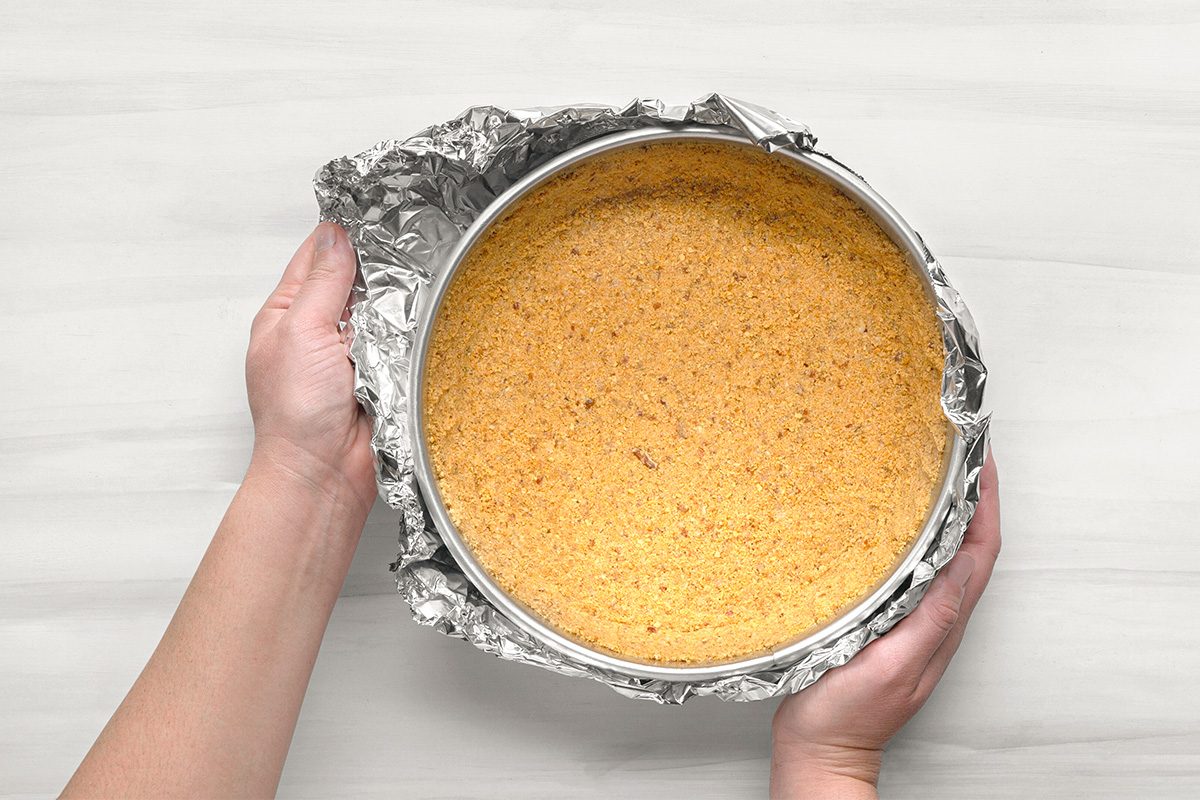 Hands holding a round springform pan lined with aluminum foil, containing a pressed graham cracker crust. The pan is placed on a light-colored surface.
