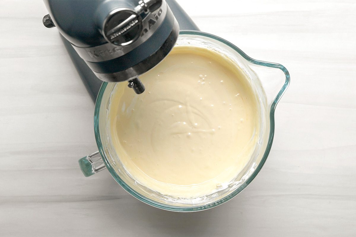 A stand mixer with a glass bowl is mixing a smooth, creamy batter on a light-colored countertop. The batter appears thick and well-blended.