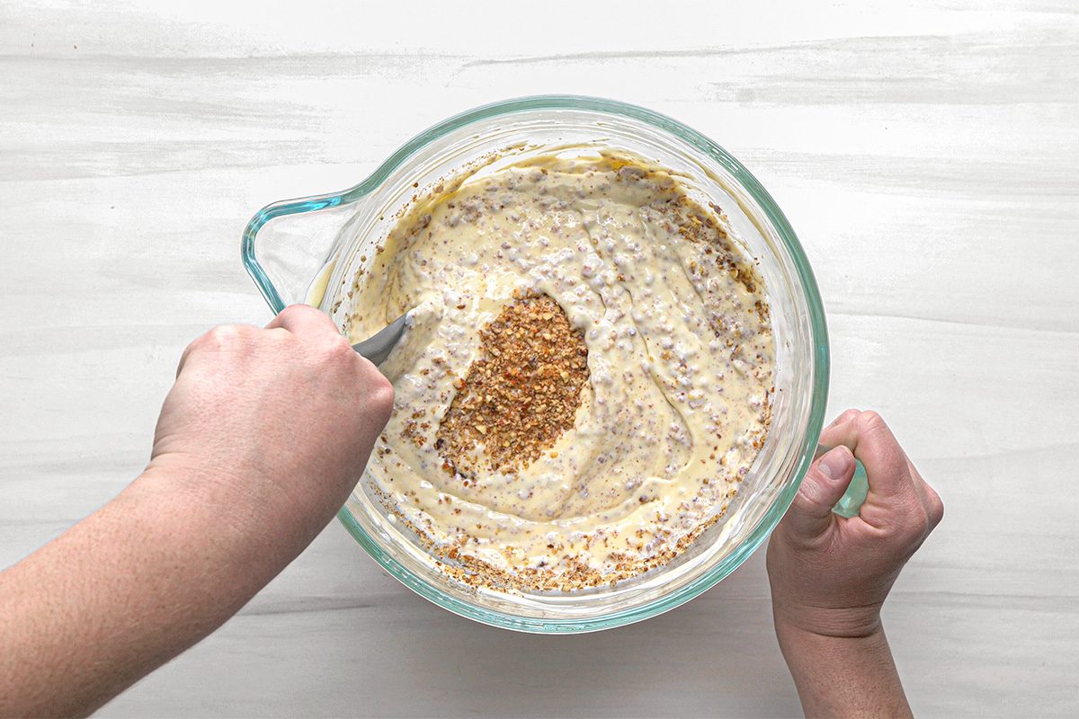 A person stirs a thick, creamy batter with chopped nuts in a clear glass mixing bowl on a white surface.