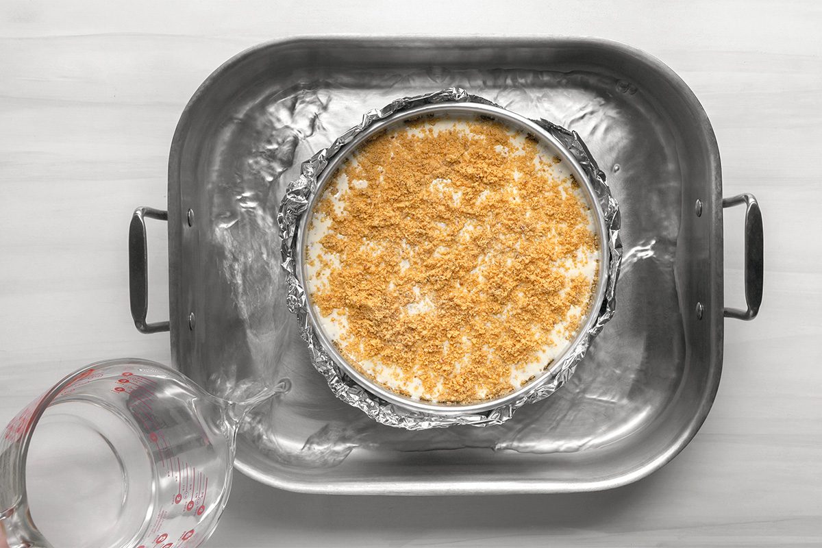 A springform pan with a crumb-topped cheesecake sits in a large roasting pan. Water is being poured from a measuring cup into the roasting pan for a water bath. The scene is on a light-colored surface.