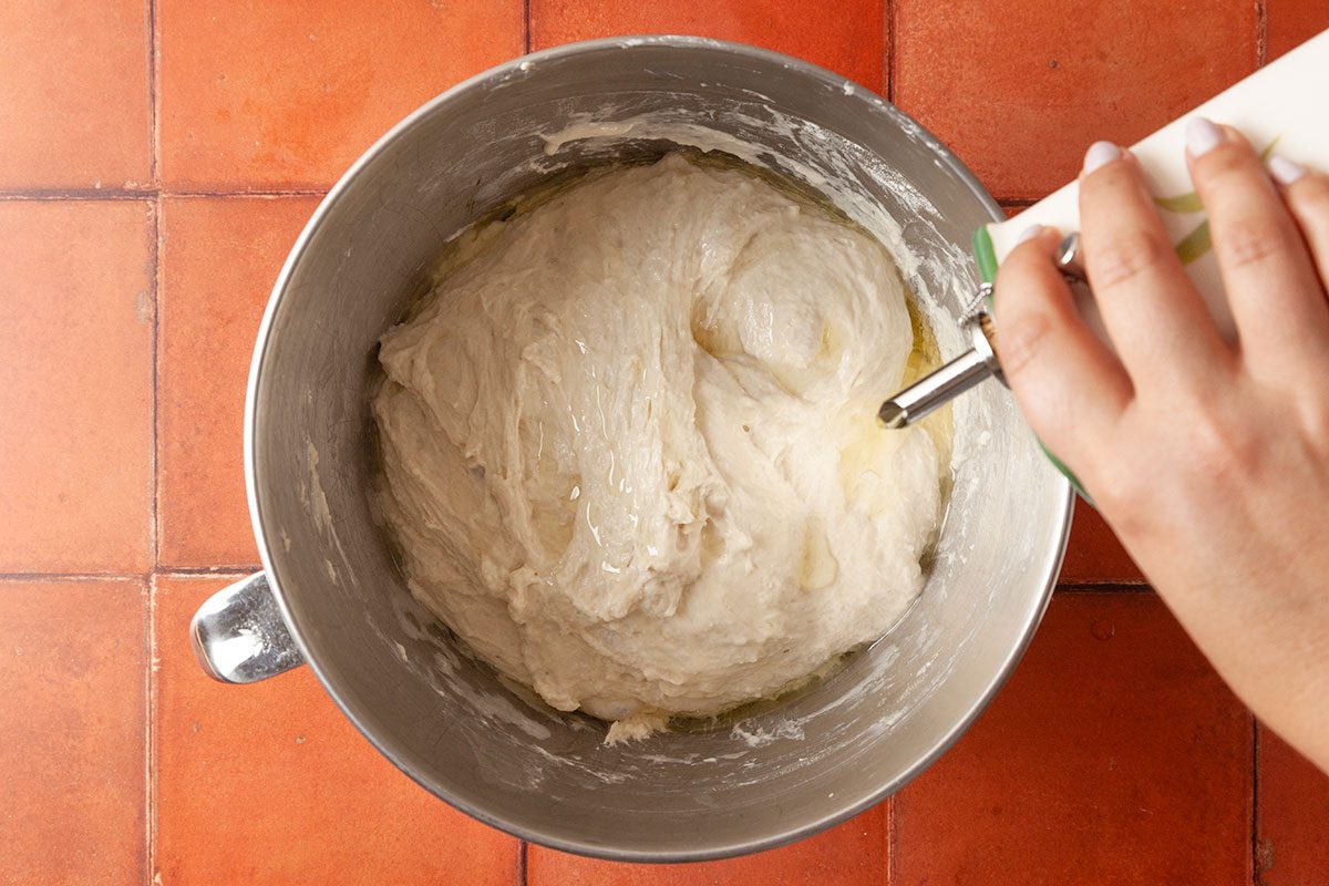 Overhead shot of a hand pouring oil from a container into a metal bowl filled with bread dough, set on a red tiled countertop.