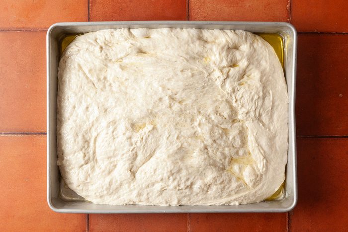Overhead shot of a rectangular baking pan filled with raw focaccia dough drizzled with olive oil, set on an orange-brown tiled surface.