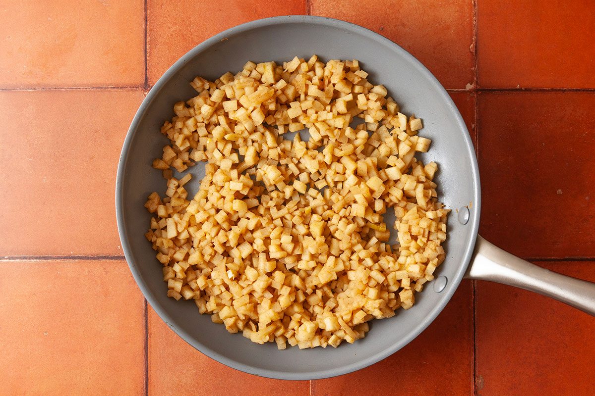 Overhead shot of golden-brown diced potatoes sautéing in a gray frying pan, set on a terracotta tiled surface.