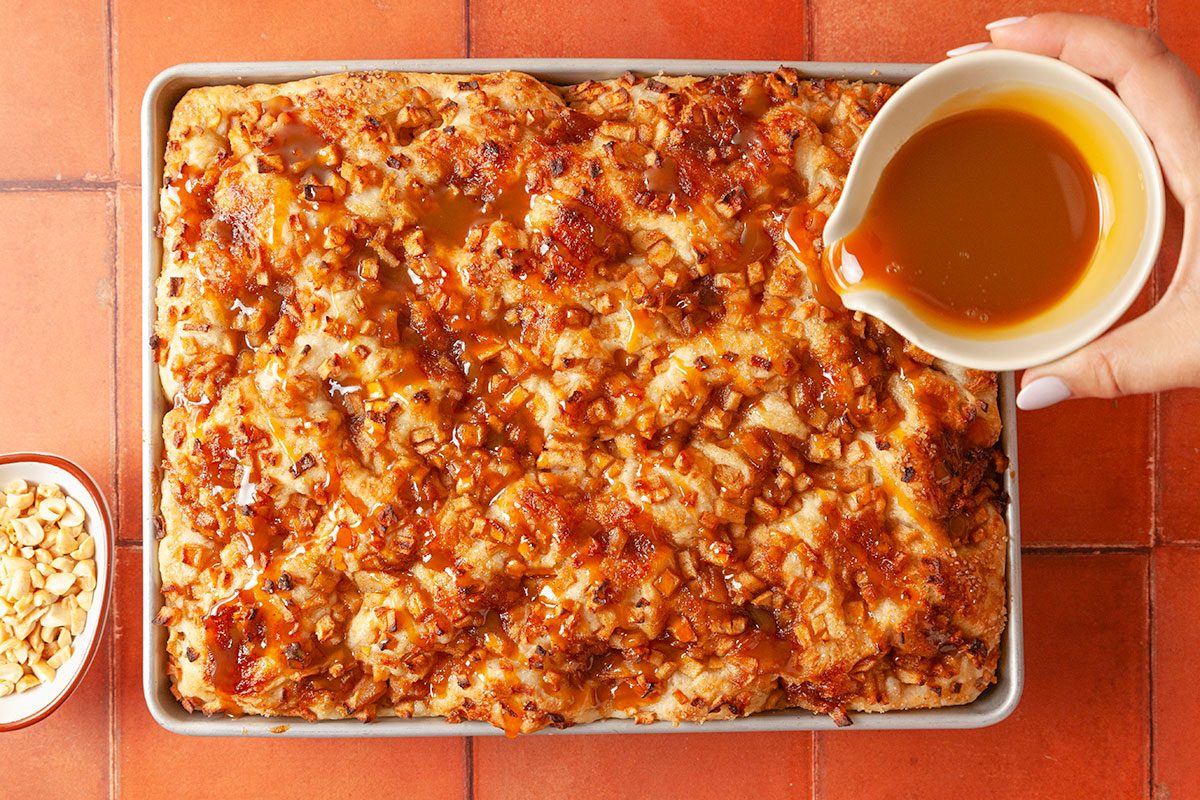 Overhead shot of a hand pouring caramel sauce over a freshly baked sheet cake topped with chopped nuts, on a tiled surface; with a small bowl of extra nuts to the side.