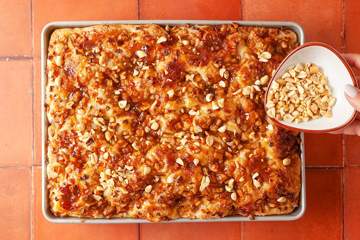 Overhead shot of a hand sprinkling chopped peanuts from a bowl onto a rectangular tray of golden baked bread or cake topped with caramelized crunchy bits; set on a reddish tiled surface.
