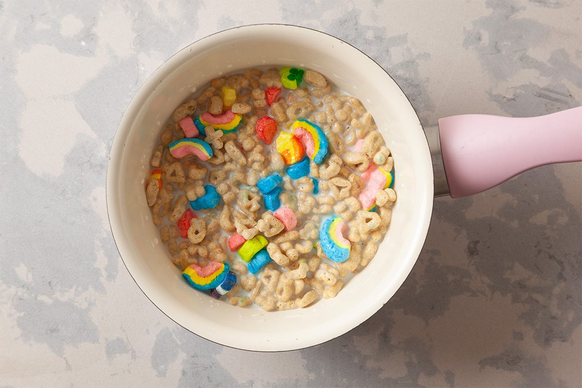A white saucepan with a pink handle contains cereal with colorful marshmallows in the shapes of rainbows, stars, and clovers, sitting on a light gray, marbled surface.