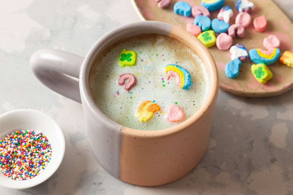 A mug of frothy milk topped with colorful marshmallow shapes and sprinkles sits next to a small bowl of rainbow sprinkles and a plate with more marshmallow shapes.