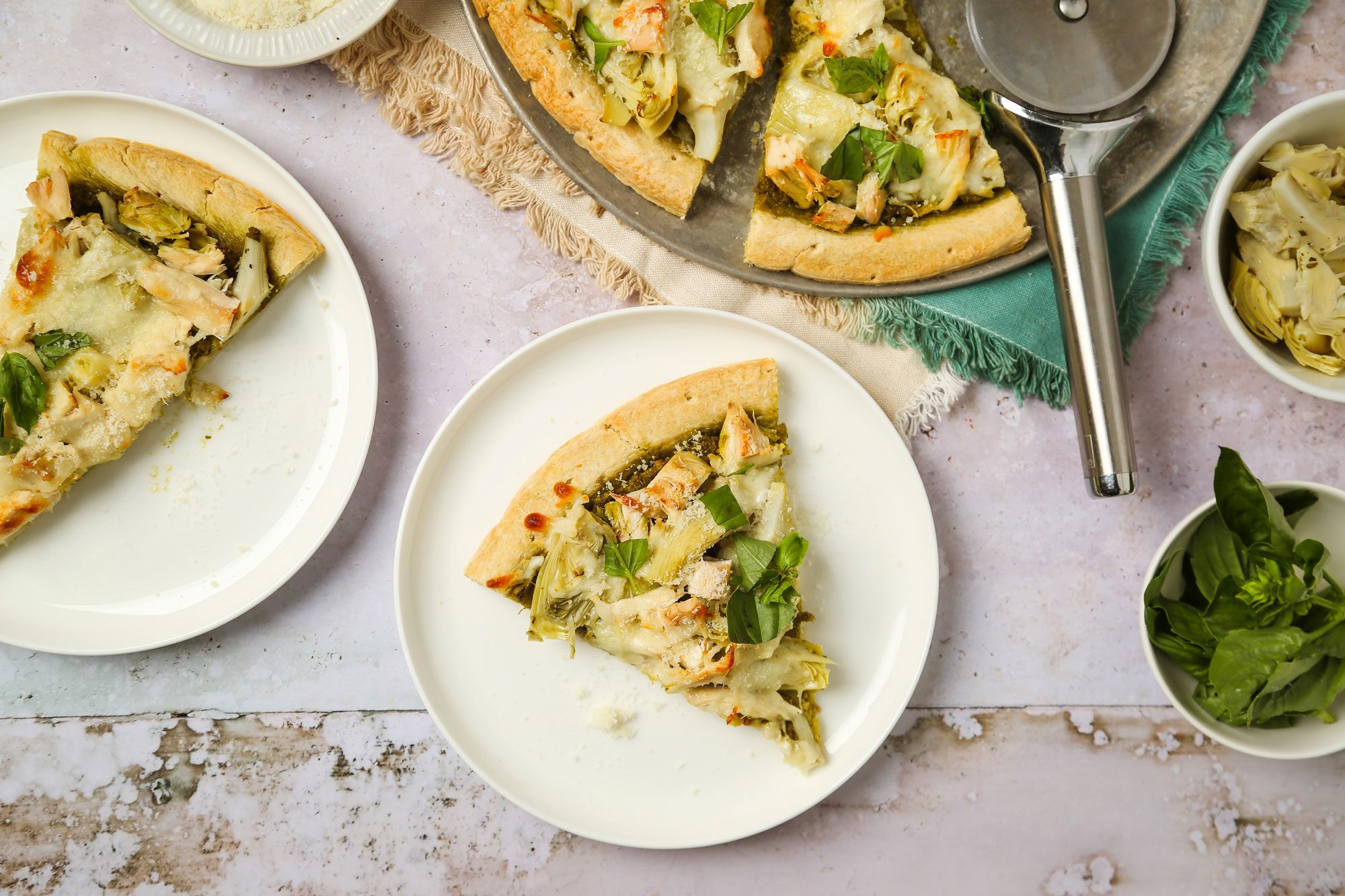 overhead shot of Two slices of vegetable pizza with melted cheese are served on white plates; a pizza cutter, a partially cut pizza, and a small bowl of fresh basil are nearby on a textured surface