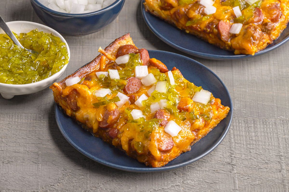 Close up shot of Chili Dog Pizza; Two square pizza slices with onions green relish sliced hot dogs and cheese sit on dark blue plates beside bowls of onions and relish