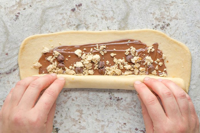 Hands roll up a strip of dough spread with chocolate and sprinkled with crumbled cookies on a light surface.
