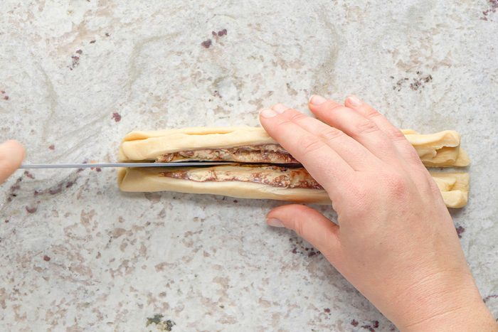 Two hands hold a rolled piece of dough with filling, while a knife cuts it lengthwise down the center on a light-colored surface.