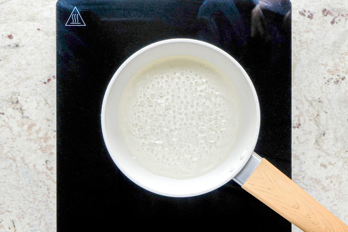 A white saucepan with a light wooden handle is heating a bubbling liquid on a black stovetop, viewed from above. The stovetop is set on a light-colored countertop.