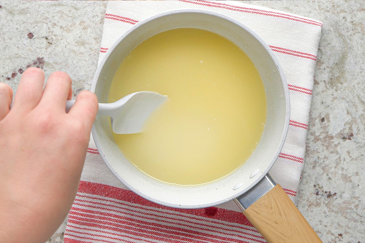 A hand stirs a pale yellow liquid in a white saucepan with a white spatula. The pan is placed on a red and white striped towel on a light-colored countertop.