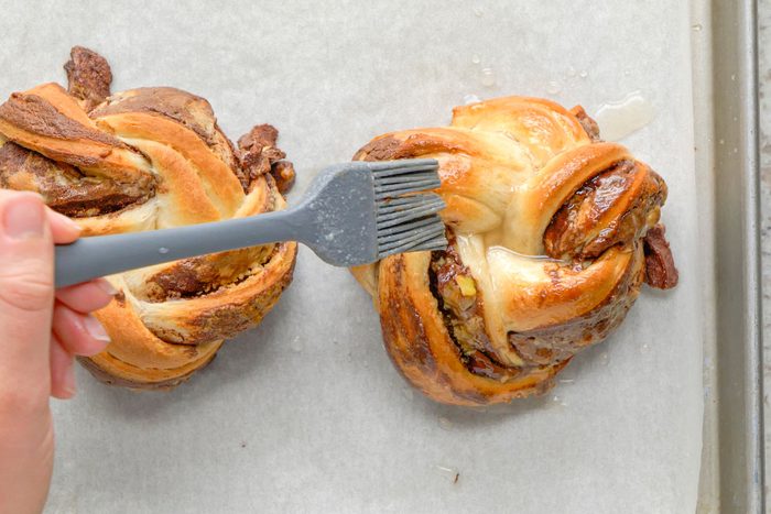 A hand uses a pastry brush to glaze two twisted, round babka buns filled with chocolate and nuts on a parchment-lined baking sheet.