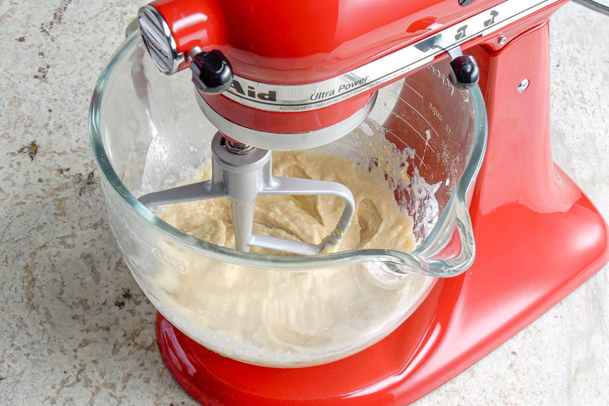 A red stand mixer with a glass bowl is mixing a creamy batter on a light-colored countertop.