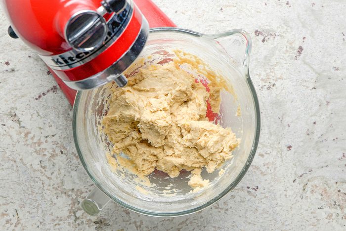 A red stand mixer with a glass bowl contains partially mixed cookie dough on a light-colored countertop.