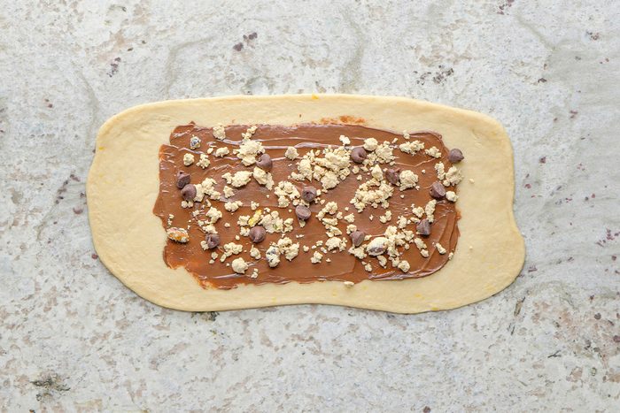 A rectangle of dough on a light countertop is spread with chocolate hazelnut spread and sprinkled with chopped nuts and chocolate chips.