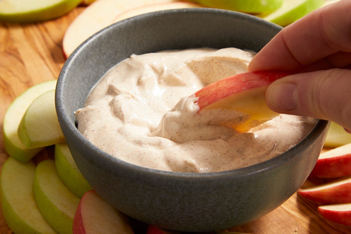 A hand dips a red apple slice into a creamy, light brown dip in a gray bowl, surrounded by green and red apple slices on a wooden surface.