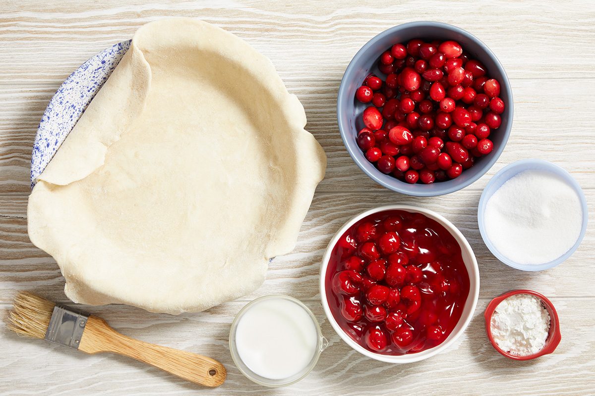 Overhead view of an unbaked pie crust in a pan, bowls of fresh cranberries, cherry pie filling, sugar, cornstarch, a cup of milk, and a pastry brush on a light-colored wooden surface.