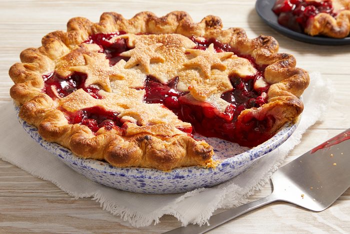 A cherry pie with a golden, star-shaped crust sits on a white cloth. One slice is missing, revealing the red cherry filling. A pie server and a plate with a slice of pie are in the background.