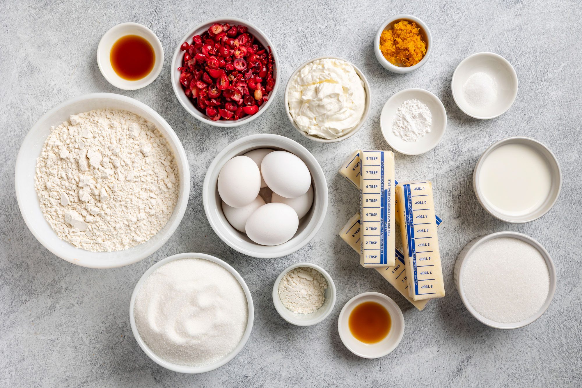 Overhead shot of various baking ingredients on a gray surface; including bowls of flour, sugar, eggs, chopped cranberries, butter sticks, sour cream, milk; and small bowls of baking powder; vanilla; orange zest and salt;