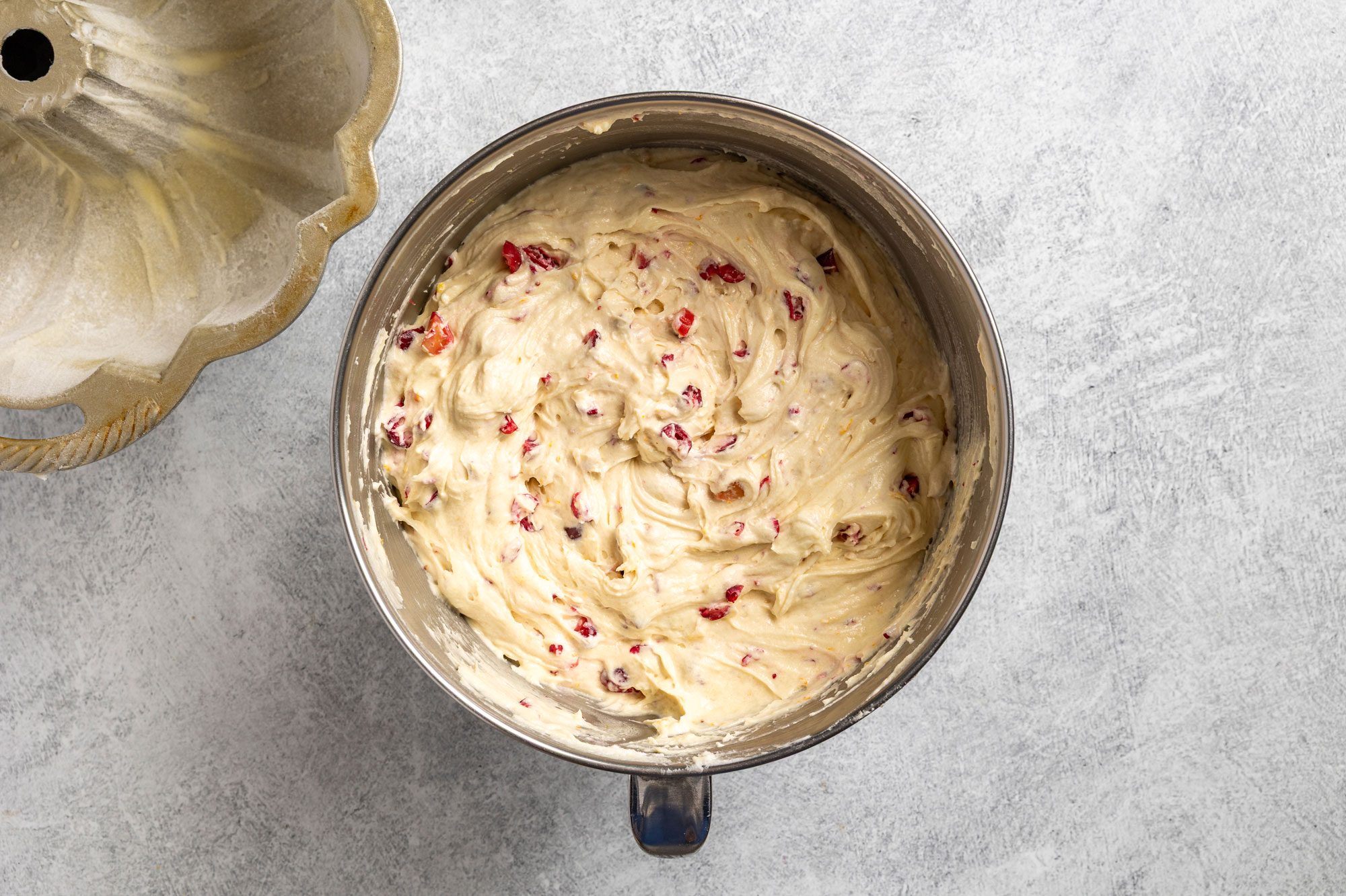 Overhead shot of a metal mixing bowl filled with creamy cake batter mixed with red fruit pieces; sitting on a gray countertop beside an empty bundt cake pan;