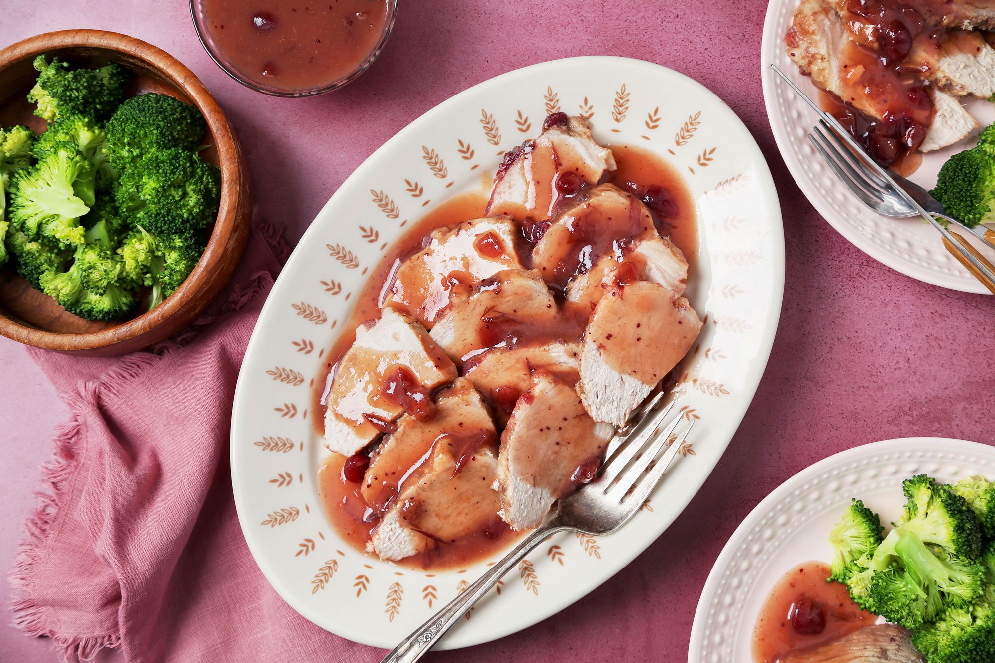 overhead shot of a plate of pork tenderloin, the pork is cut into slices and covered in a red cranberry sauce, a fork is placed on the side of the plate, on the left side of the image there is a bowl of broccoli with a pink cloth next to it, on the right side there are two plates with broccoli, one plate has a slice of pork with cranberry sauce; the image has a pink background