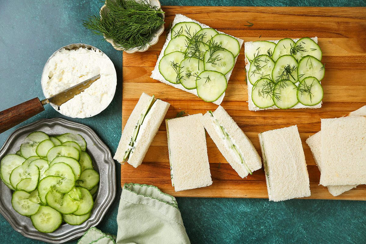 A wooden board with cucumber tea sandwiches, some topped with cucumber slices and dill, others cut into rectangles. Nearby are a bowl of sliced cucumbers, a dish of cream cheese, a knife, and a sprig of dill.
