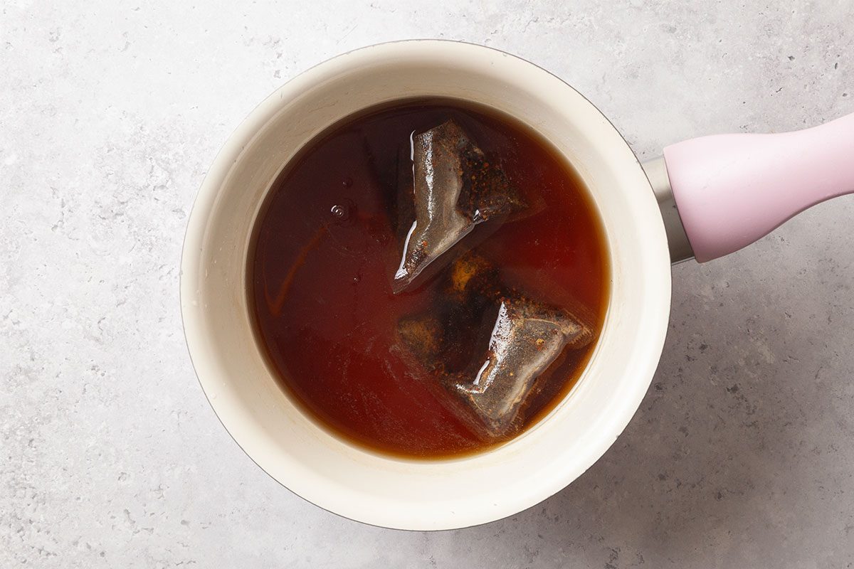A white saucepan with a pink handle contains hot water and two tea bags steeping, creating a dark brown liquid, set on a light gray surface.