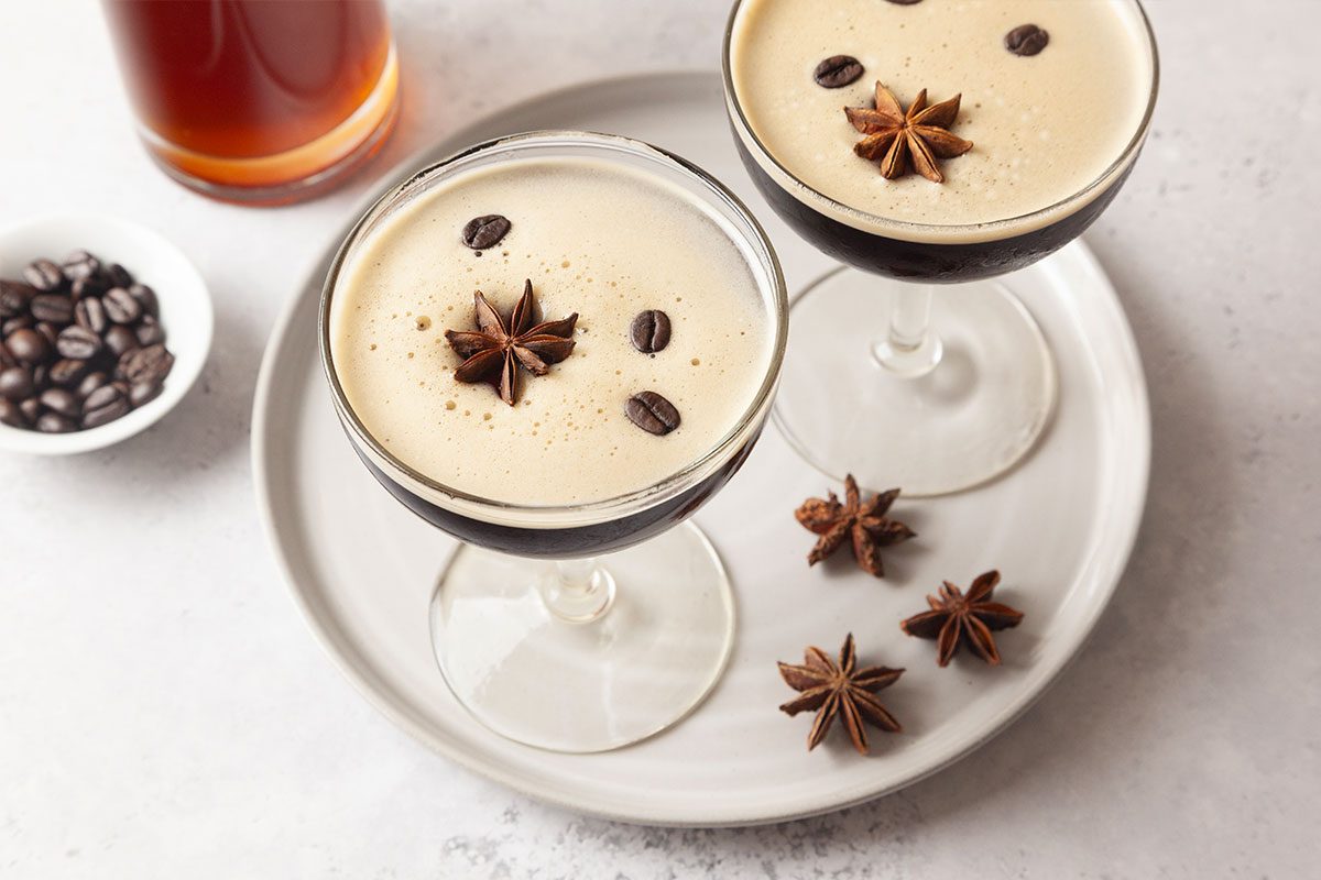 Two espresso martinis in coupe glasses on a white plate, each garnished with star anise and coffee beans. A small bowl of coffee beans and a bottle are in the background.
