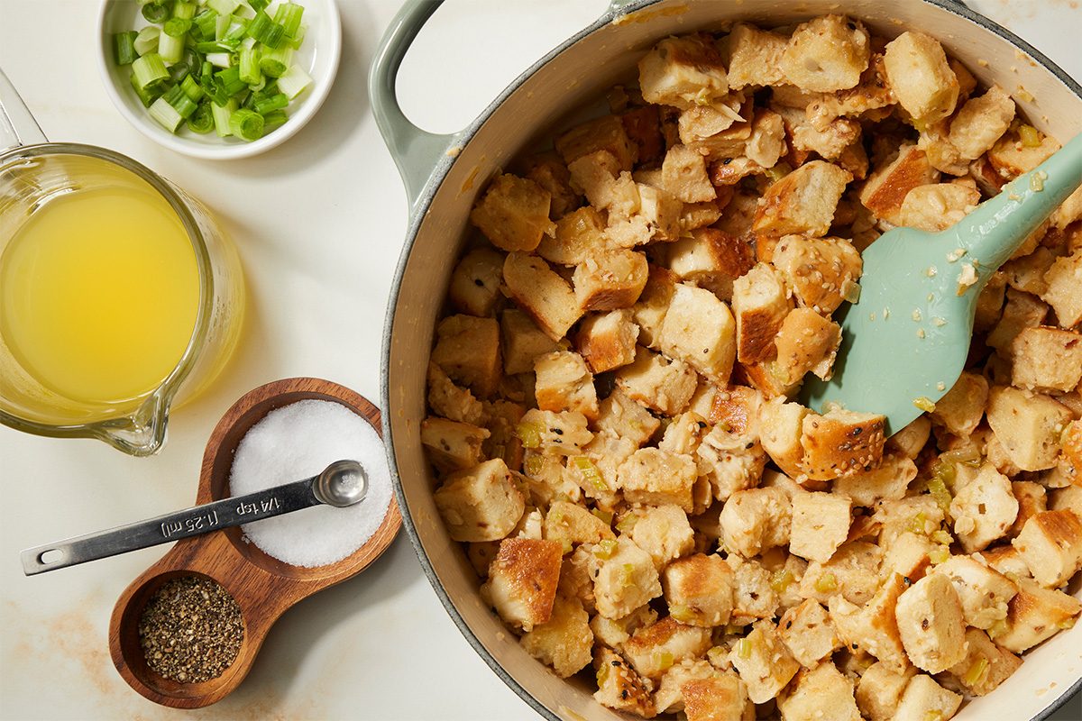 A pot of bread cubes mixed with sautéed vegetables, next to a bowl of chopped green onions, a measuring cup of broth, and containers of salt and pepper on a white countertop.